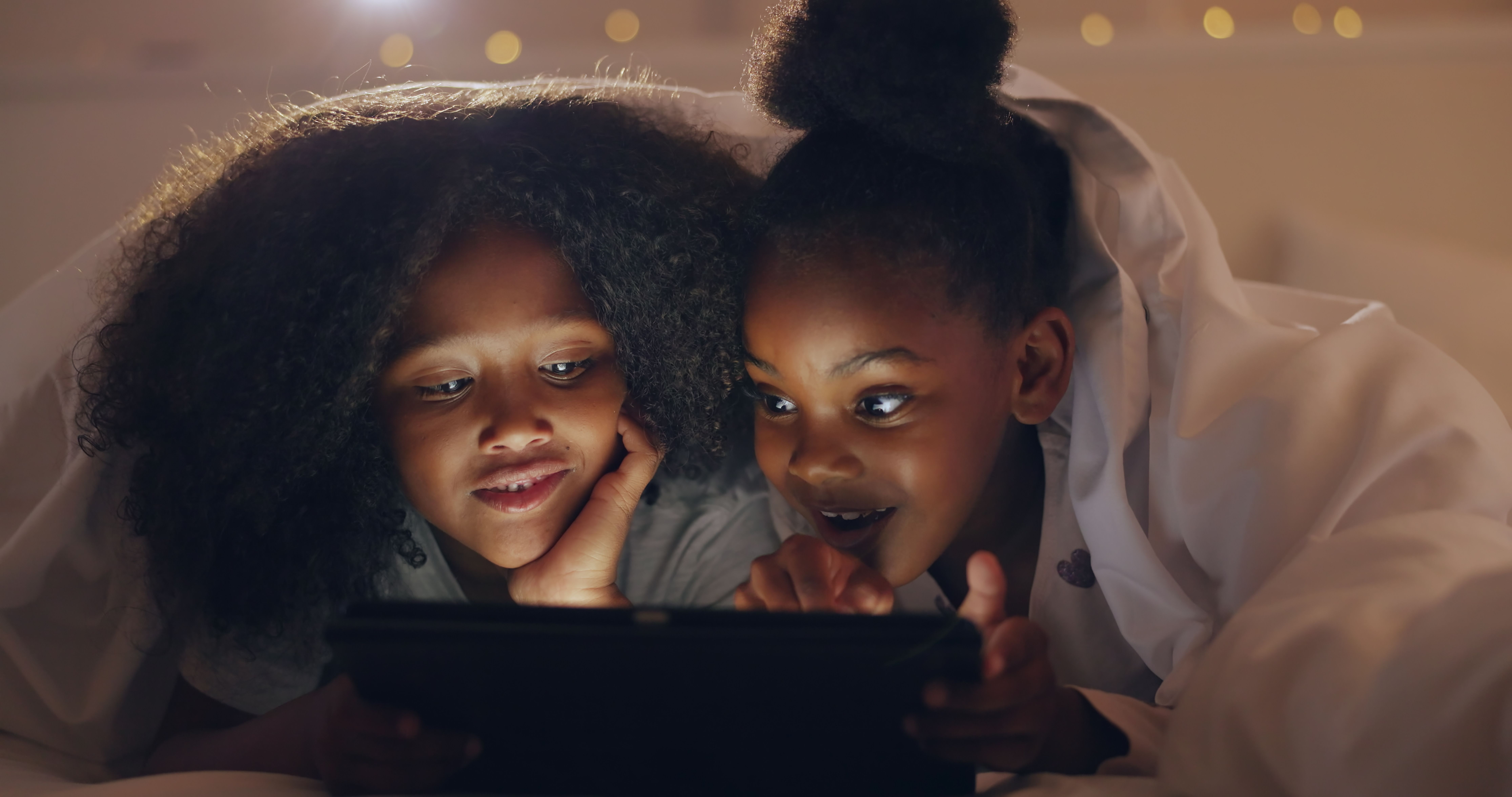 Sisters under a blanket, faces lit by tablet glow